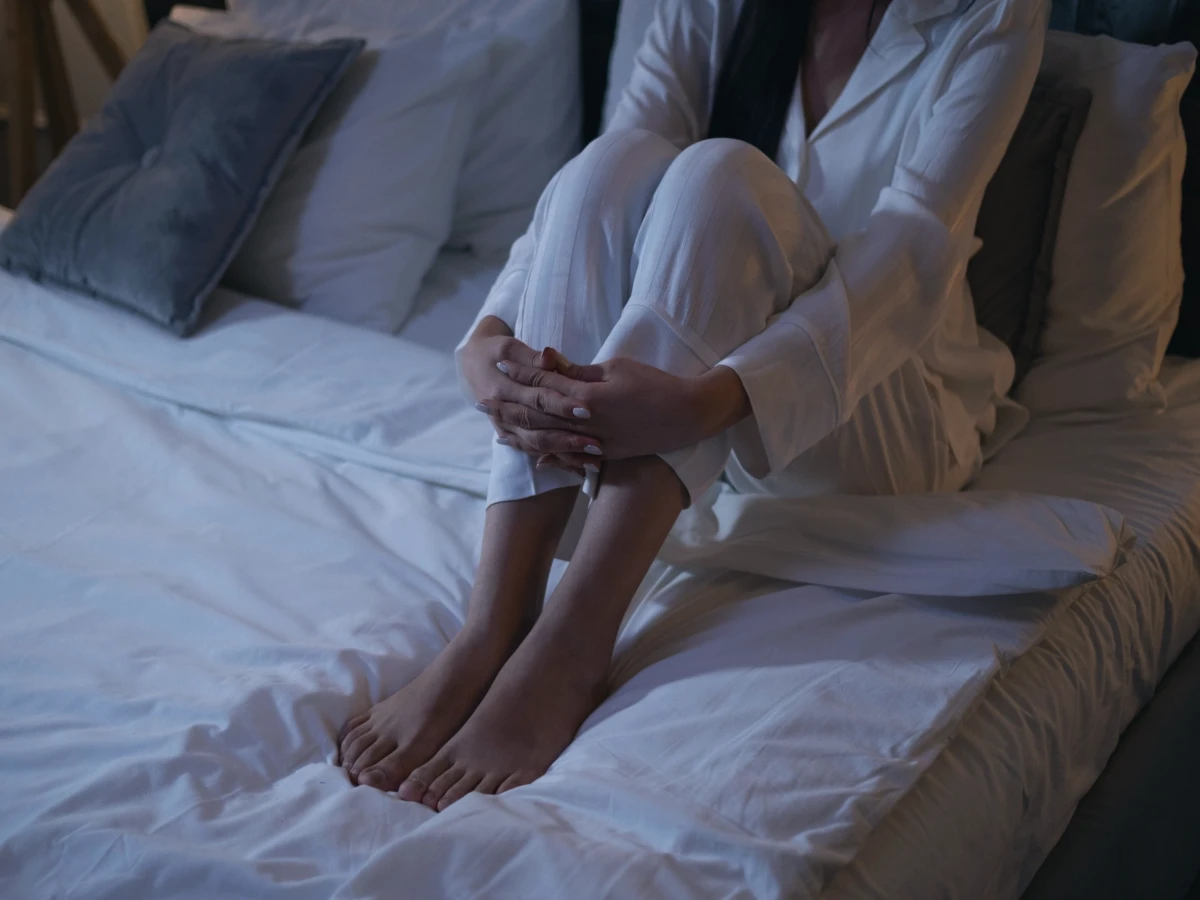 A person sitting on the edge of a bed at night with bare feet on the floor