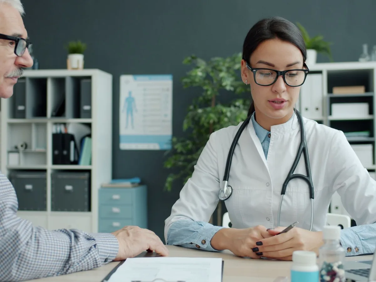 A clinician talking with an older man at a desk, with medication bottles and notes in front of them