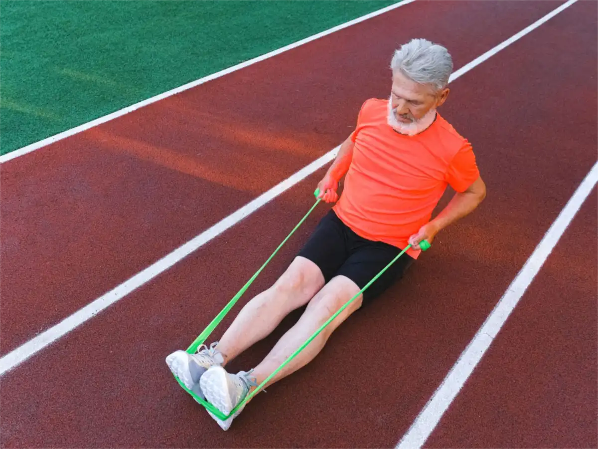 An older man sitting on a running track with a green resistance band looped around both sneakers