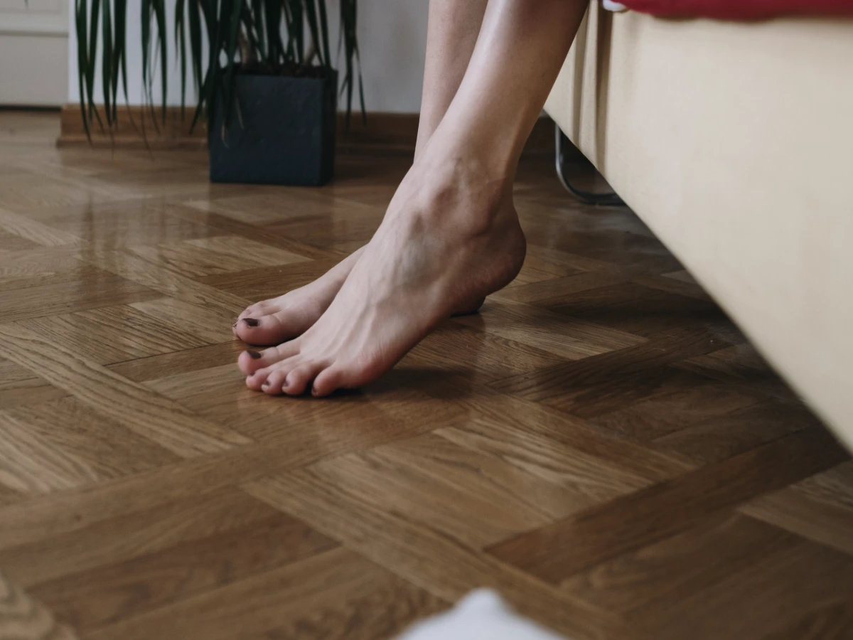 Bare feet resting on a wooden bedroom floor beside a bed in morning light