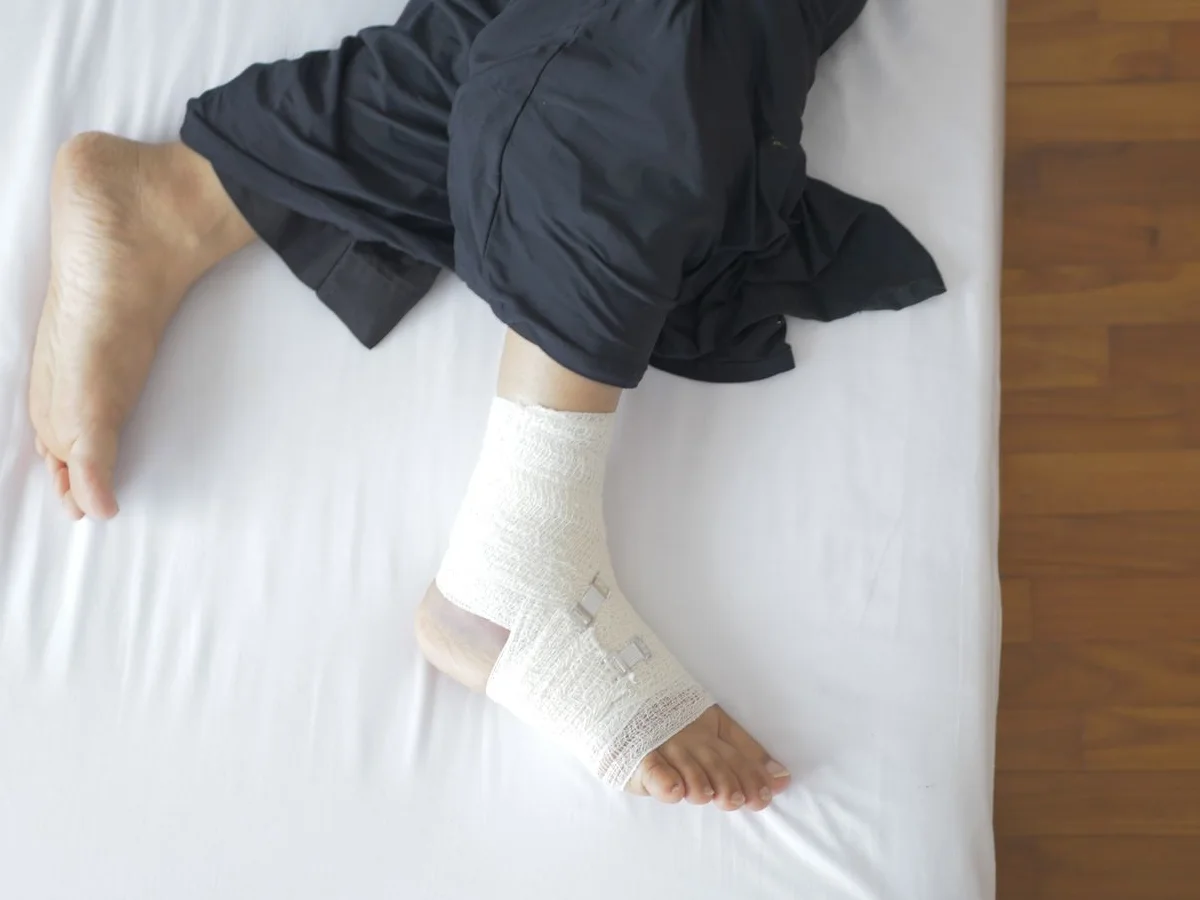 A foot and ankle wrapped in a clean white bandage, resting on a bed beside the other bare foot