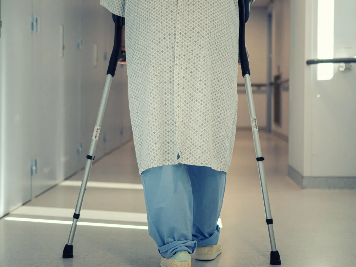 A patient in a hospital gown walking down a hospital corridor on crutches, seen from the chest down