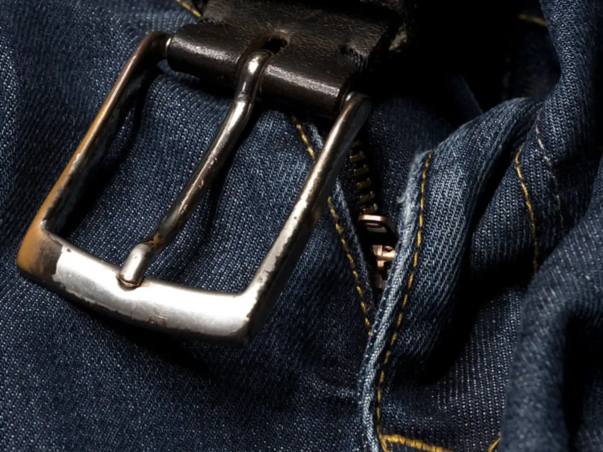 Close-up of a metal belt buckle and the zipper of a pair of dark blue jeans