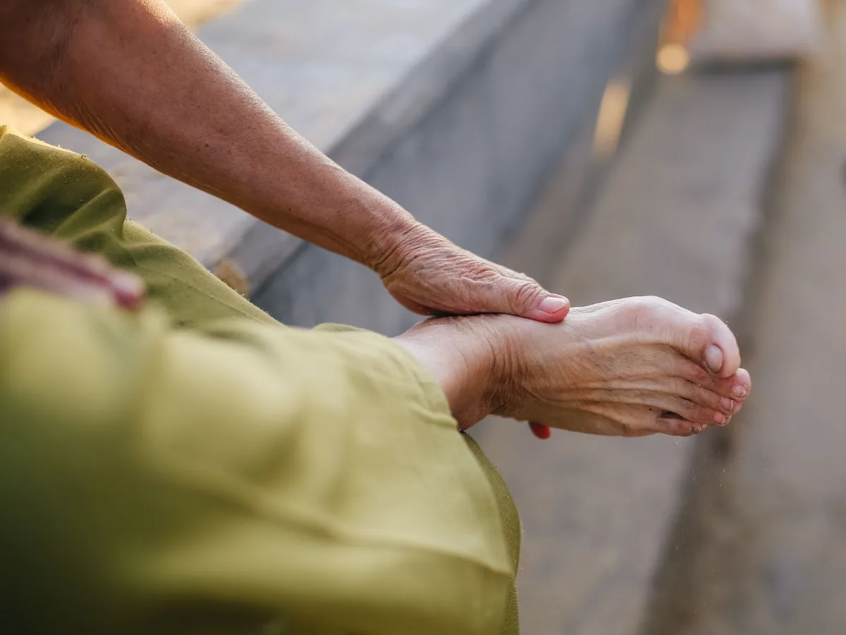 An older person sitting outdoors, holding the top of one bare foot